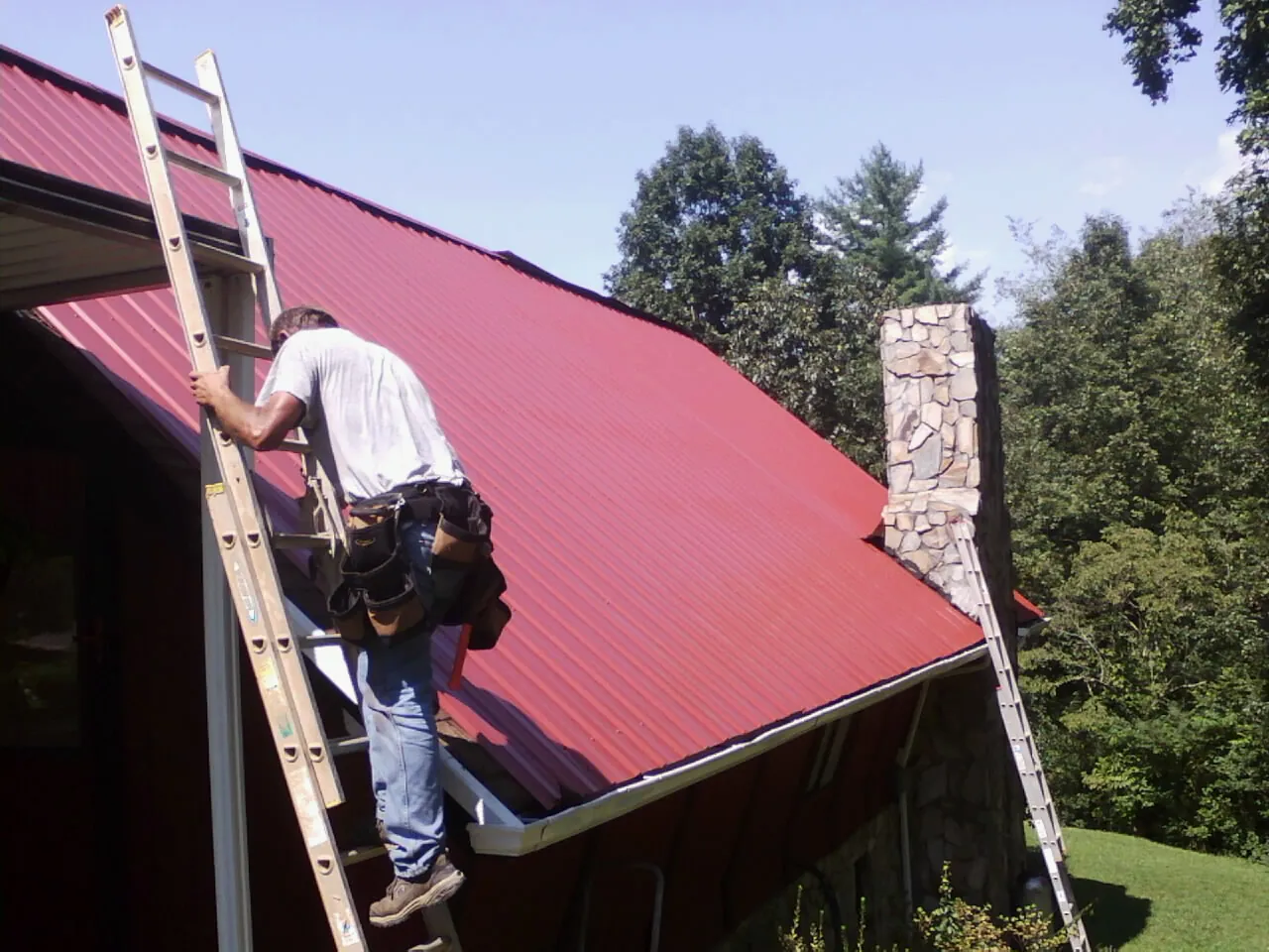 Roofer on a ladder beside a red metal roof and stone chimney — Soffit & Fascia Repair by Roofing Brothers in Sun City Center